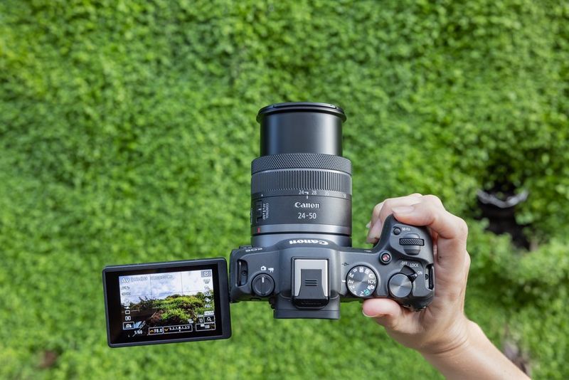 Een hand houdt een Canon EOS R8-camera vast. We zien de bovenkant van de camera met het objectief uitgeschoven en het variangle-touchscreen uitgevouwen.