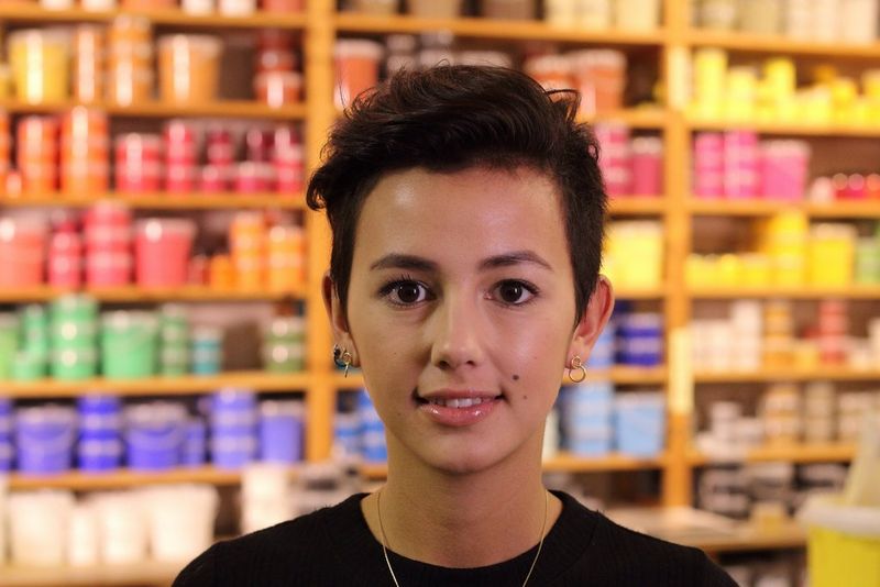 A portrait of a young woman against a background of shelves filled with colourful items, still blurred but more discernible.