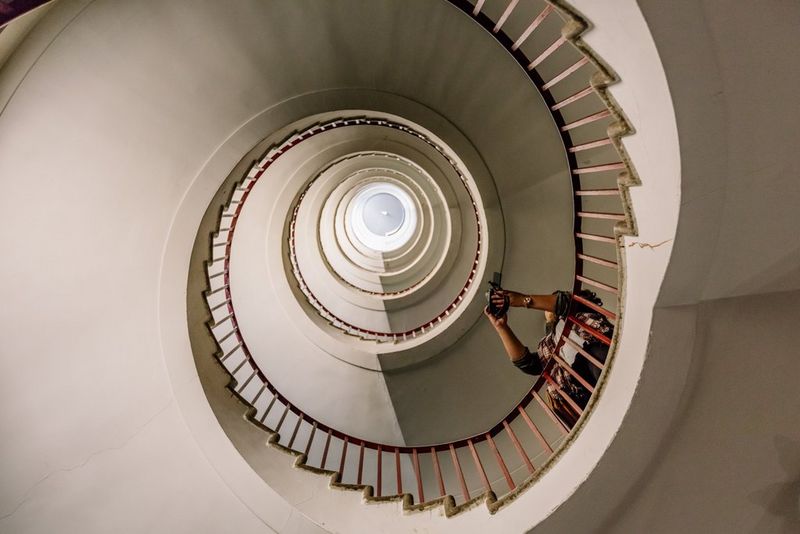 תמונה שצולמה כלפי מעלה מתוך גרם מדרגות לולייניות מבטון היוצר דוגמה מפותלת דמוית קונכייה, בזמן שגבר העומד על גרם המדרגות מכוון מצלמה כלפי מעלה.