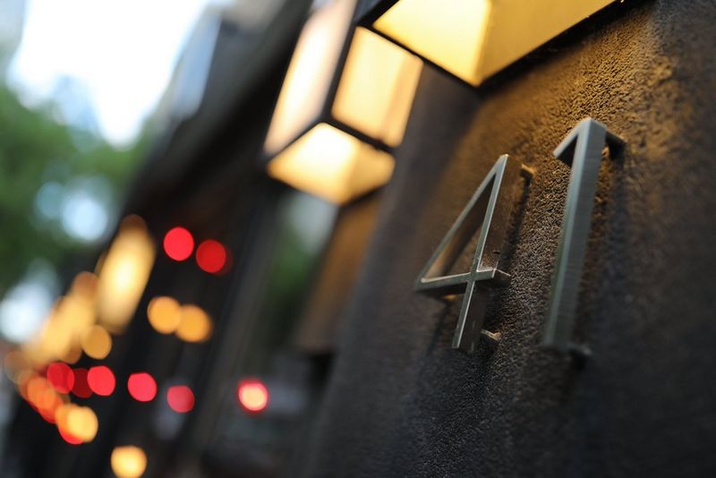 Eine Nahaufnahme einer Gebäudeseite, eine Metallhausnummer, die von der Wandleuchte darüber beleuchtet wird.