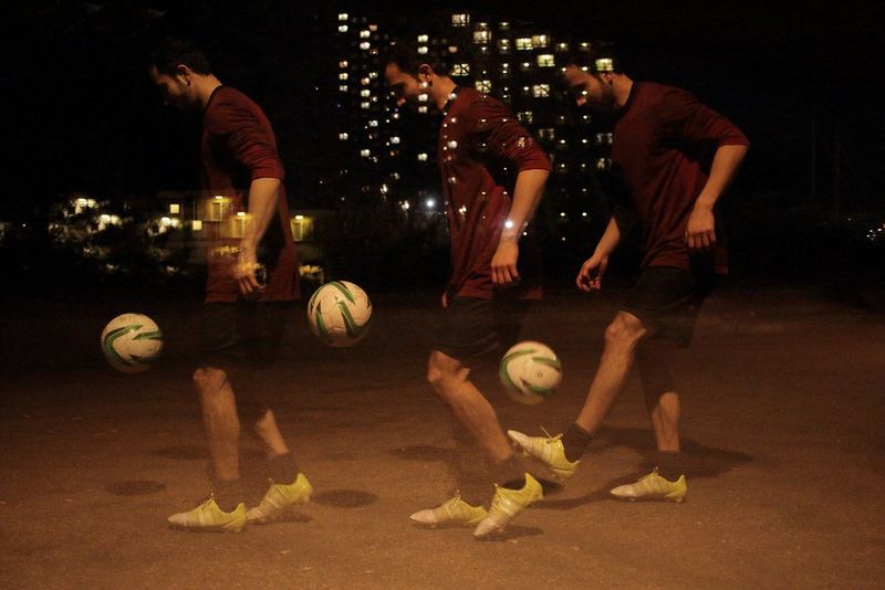 Uno scatto a esposizione multipla di un uomo in pantaloncini e maglietta con un pallone da calcio di sera, con le luci degli edifici sullo sfondo.