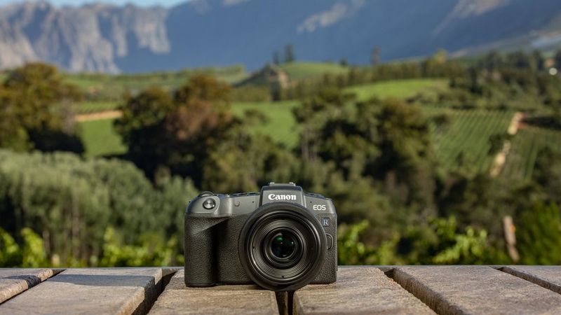 A Canon EOS RP camera is positioned on a wooden table with rolling green landscape behind it.