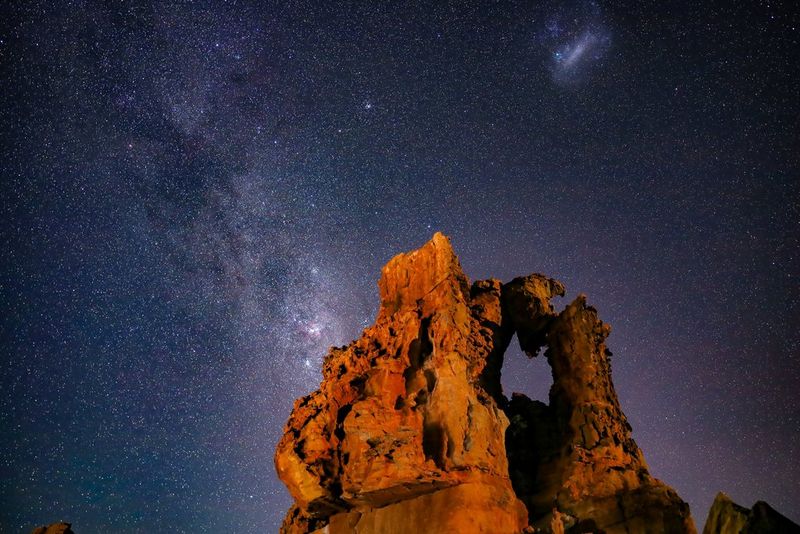 A starry night sky with an orange rock formation in the foreground.