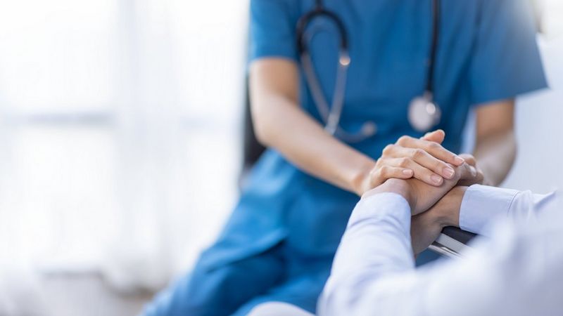 A healthcare professional in blue scrubs gently holding the hands of a seated person, offering comfort and support. A stethoscope is visible around the professional’s neck, suggesting a medical or clinical setting.