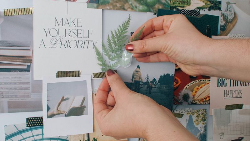 A pair of hands affixes a paper leaf to a pin board covered in notes, photos and other assorted printed items.