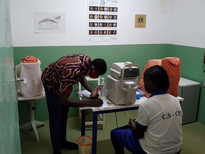 Two people in a green and white walled medical examination room. One sits in front of a machine, ready for an eye examination, the other is bent over and making notes.