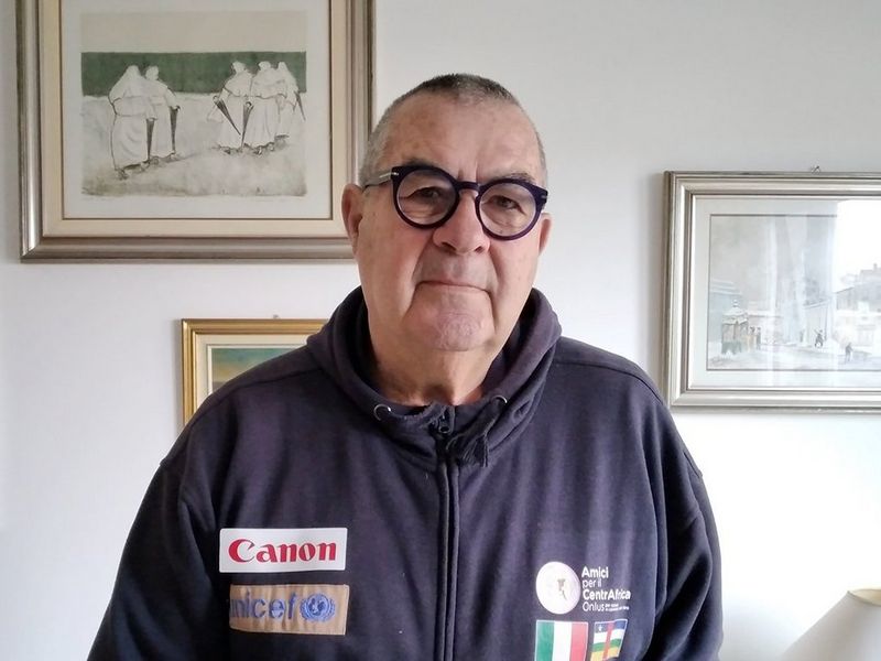 A man stands in front of a white wall, where three pictures are hanging. He wears black glasses and a navy-blue zip-up top, covered in logos, including UNICEF and Canon.