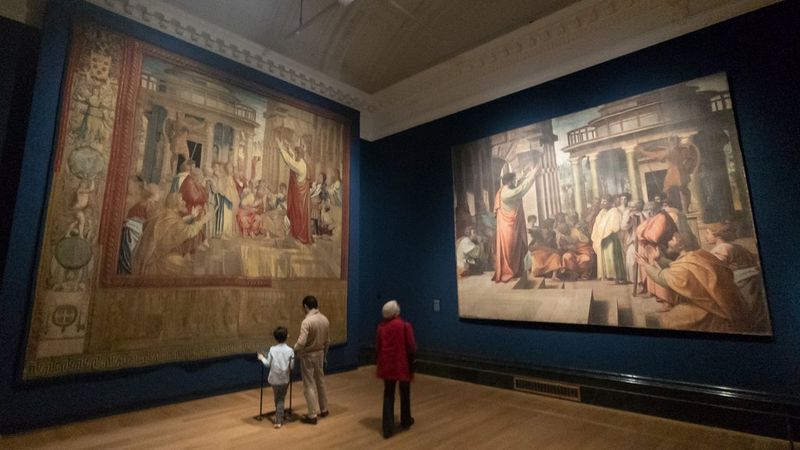 Visitors looking at large artwork reproductions hanging on dark blue walls in a gallery room.