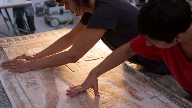 Two conservators carefully aligning and pressing down a textured print featuring ancient Egyptian motifs.