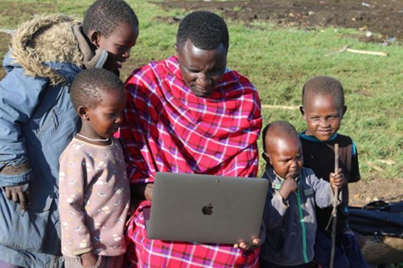 Manja holds a laptop while a group of four small children gather around to look at the screen.