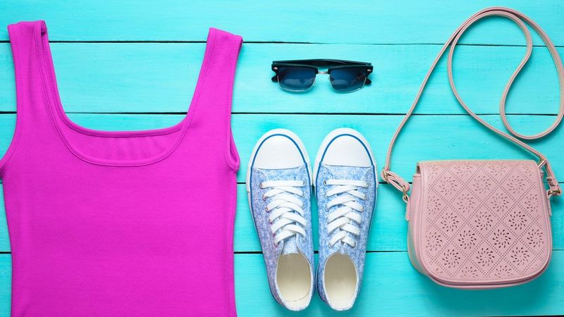 Clothing and accessories laid flat against a surface of bright blue painted wood. A violet vest, a pair of sunglasses, some blue floral converse-style plimsolls and a light pink shoulder bag with laser cut repeating patterns on the front flap.