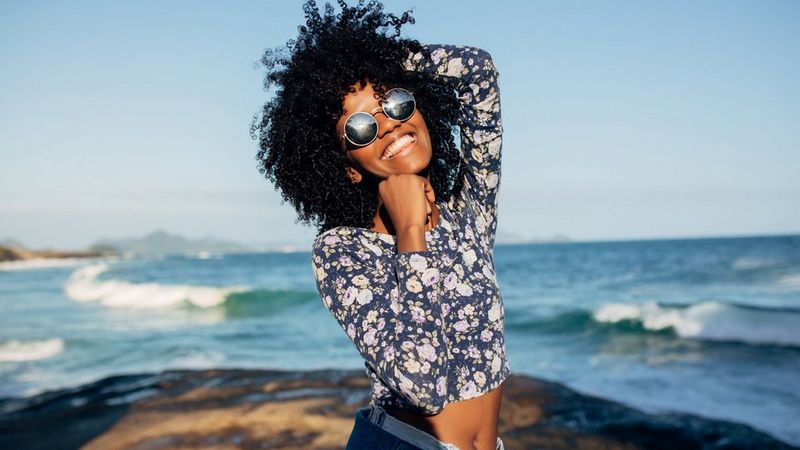 A young woman in sunglasses stands in front of a choppy sea in the sunshine. She smiles and poses for the camera. She wears a floral cropped t-shirt and jeans, with her stomach exposes. 