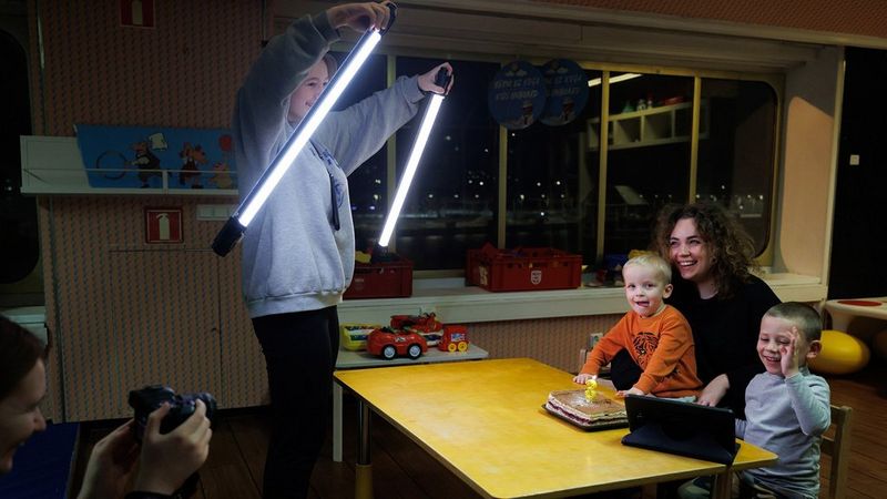 A teenage girl holds LED light tubes in front of a woman and two small children who are sat at a table. Behind her, a woman holds a camera to photograph them.