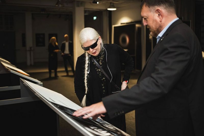 A man and a woman in smart dress appear to be looking at images and text on a plinth in front of them. The man’s left-hand rests on the plinth. The woman has long white hair, styled in a plait, and wears dark glasses