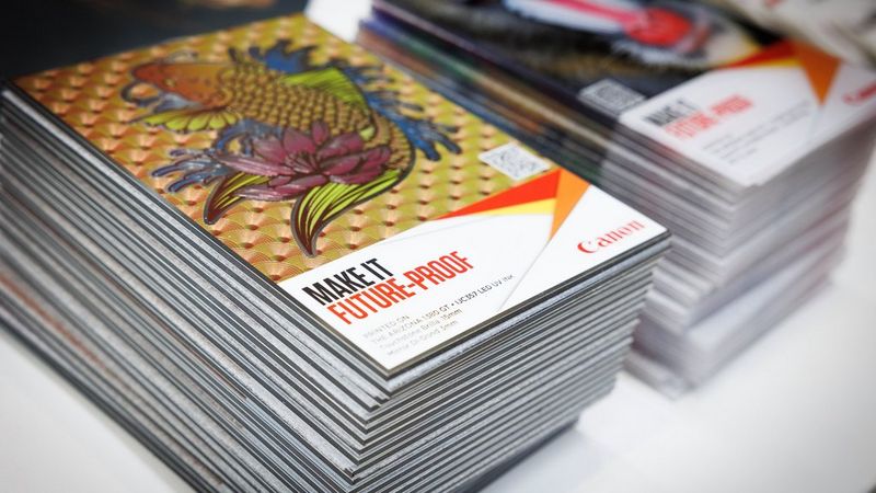 Two stacks of brochures, side by side, photographed at a slight angle from above, so you can clearly see the stack and the cover. On the cover is a Japanese-style illustration of a fish, surrounded by flowers and leaves. It is printed against a patterned background of fans. The text beneath it, in capital letters, reads, ‘make it futureproof’. Beside this text if a red, yellow and orange origami-style shape and beneath it is the red Canon logo.
