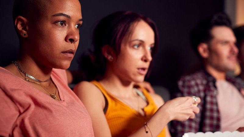 Three people sat side by side. The expressions on their faces suggest they are watching something gripping or about to be scary. The person in the middle’s hand hovers over a popcorn carton.