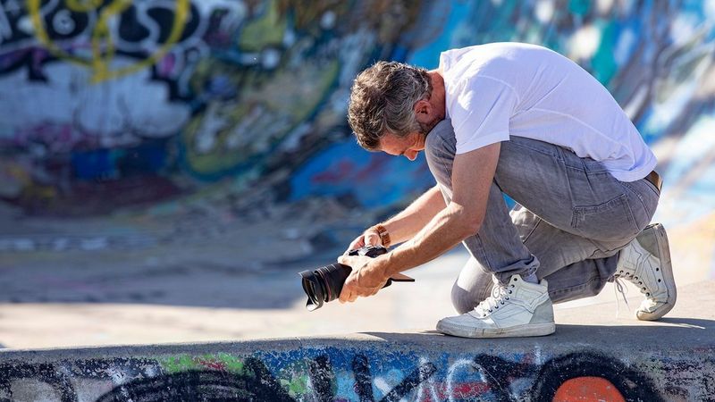A man in a white t-shirt and jeans kneels in a graffiti-covered concrete space. He holds a Canon camera and points it at the ground.