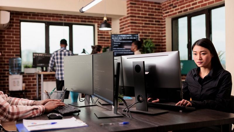 Two people sit at their desk in an office, facing each other but both looking at big screens as they work on their computers.