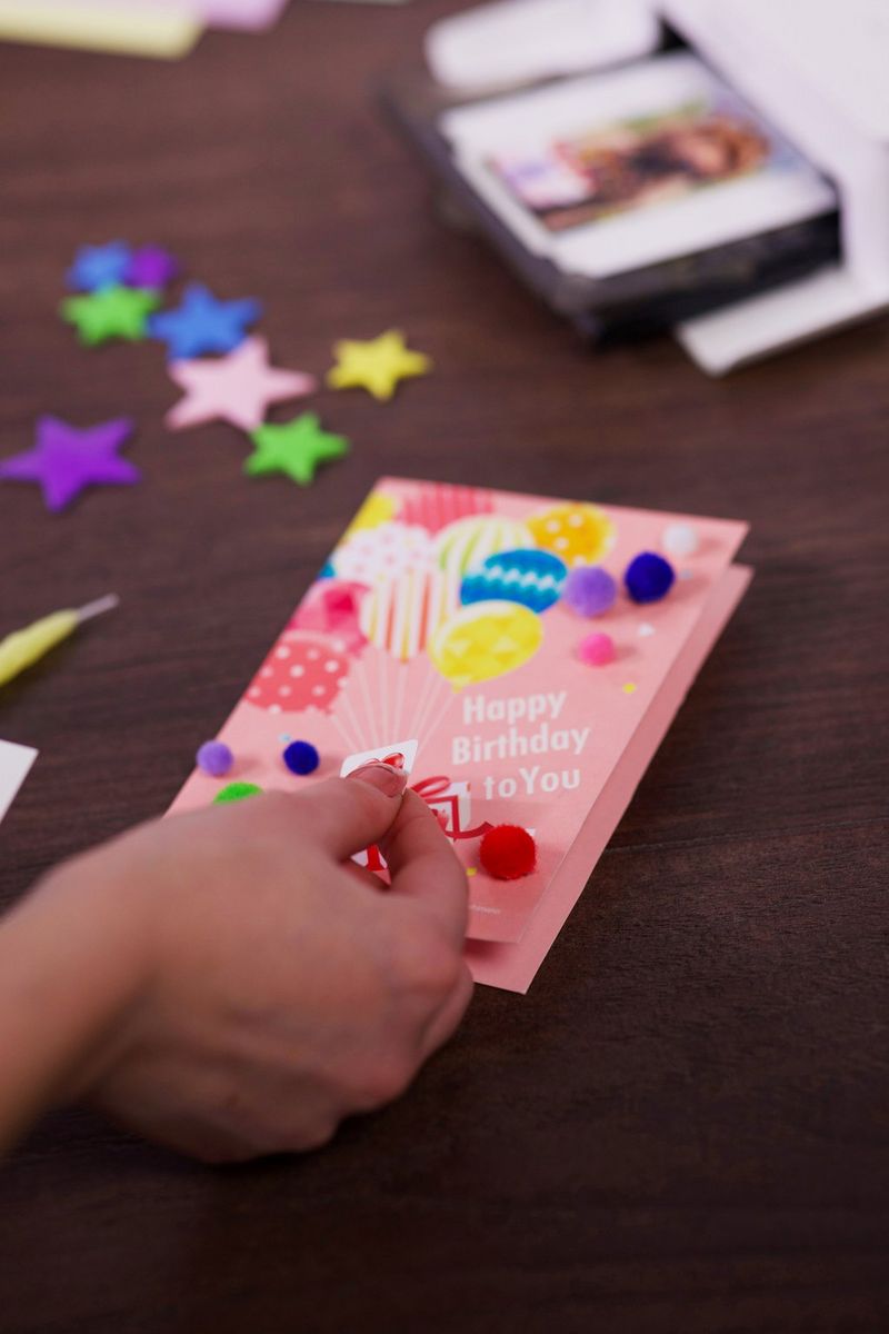 יד מוסיפה אלמנטים קישוטיים כמו פונפונים צבעוניים לכרטיס יום הולדת ורוד. מדפסת SELPHY CP1500 נראית ברקע.