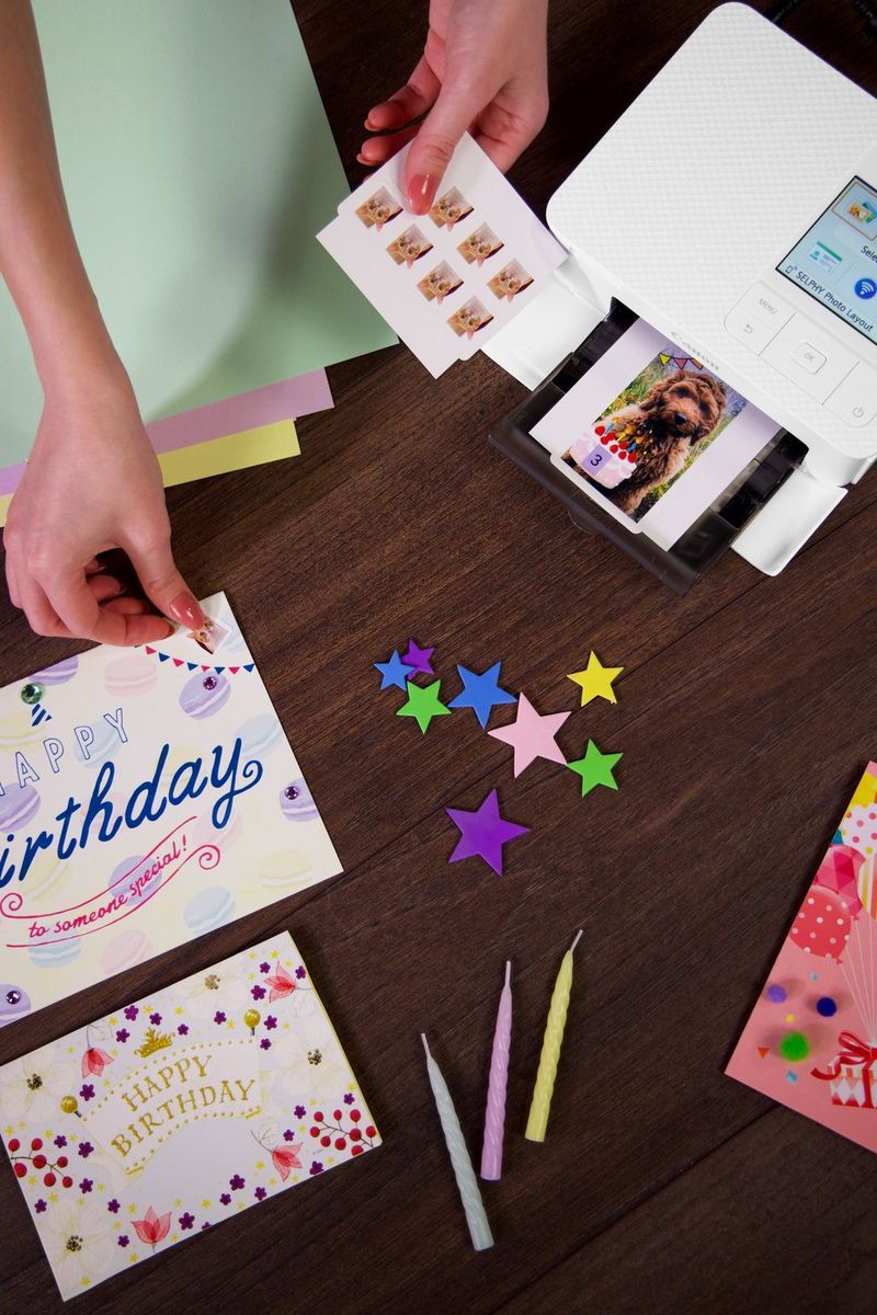יד מניחה גיליון של סטיקרים קטנים בנושא חתולים לצד כרטיס "מזל טוב", עם מדפסת SELPHY שמדפיסה תמונה של כלב עם עוגת יום הולדת לצידו.