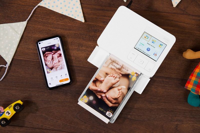 מדפסת Canon SELPHY על שולחן מעץ, לצד טלפון חכם. המדפסת מדפיסה תמונה מצולמת של כפות רגליים של תינוק.