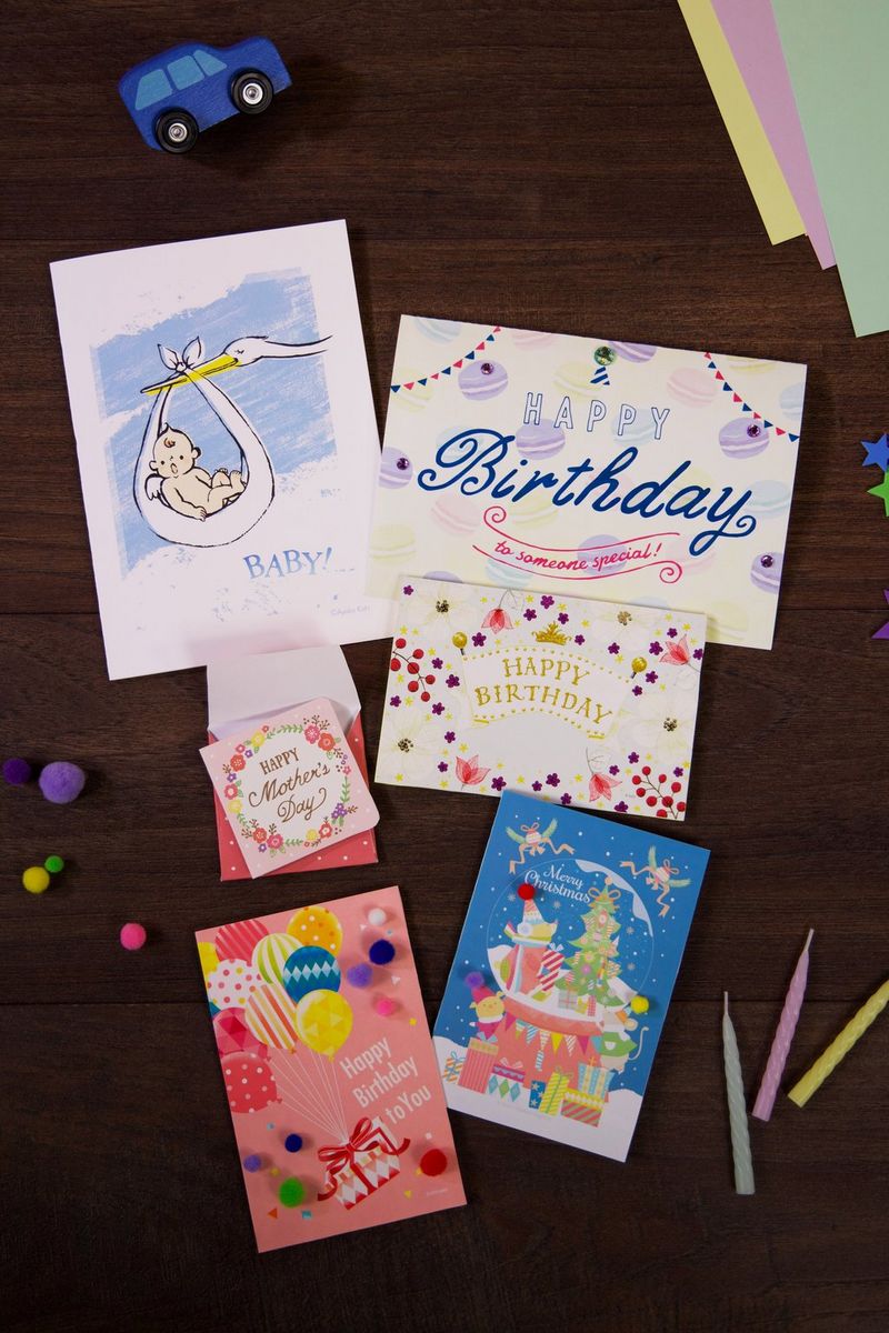 An array of celebration cards laid out on a wooden table, including a birthday card, Mother's Day card, Christmas card, and a birth announcement card, accompanied by pastel-coloured candles and bright crafting pom-poms. 