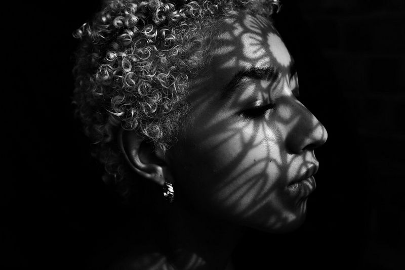 In this black and white image, a model's head is tightly framed against a black background, the shadow of a mandala design falling onto her forehead and across the rest of her face.