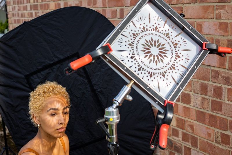 A model sits in sunlight against a black backdrop, a mandala pattern held in front of her by a tripod casting shadows across her skin.