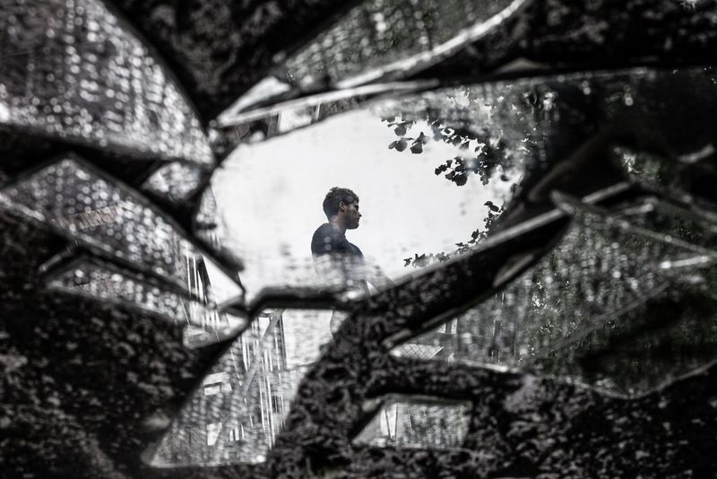 A group of mirror shards lie on the ground, the largest piece reflecting the head and torso of a man looking away.