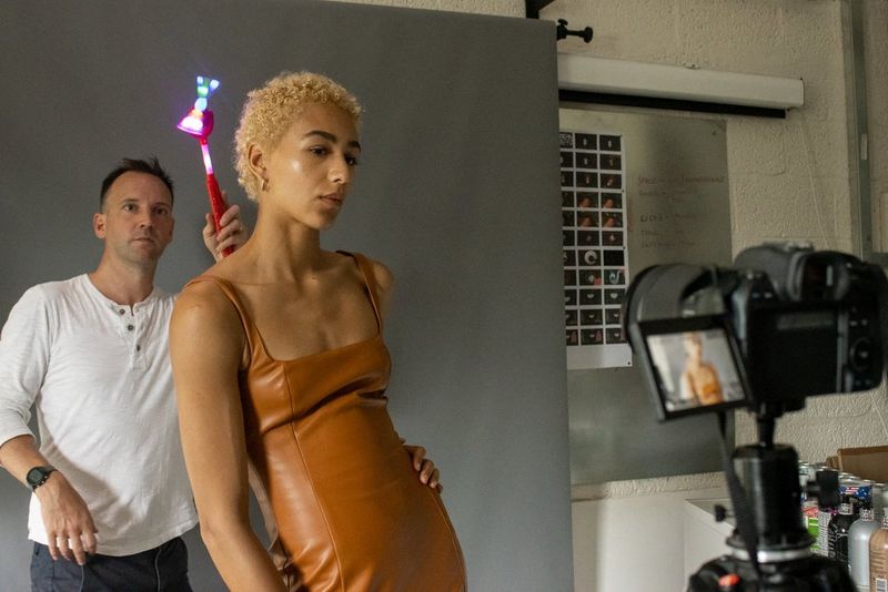 A model stands in front of a camera and tripod with one hand on her hip. Behind her, a man holds up a twirling, multi-coloured LED light.