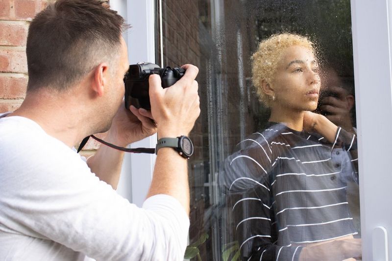 A man holds a camera up to his eye, shooting through a glass door sprinkled with water droplets to a model on the other side.