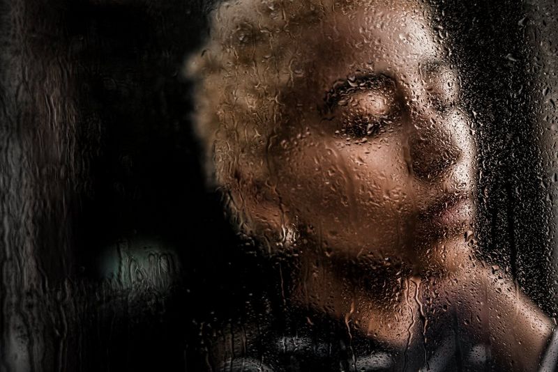 A woman looks to the side with her eyes closed. She is out of focus and viewed through glass with water droplets trickling down.