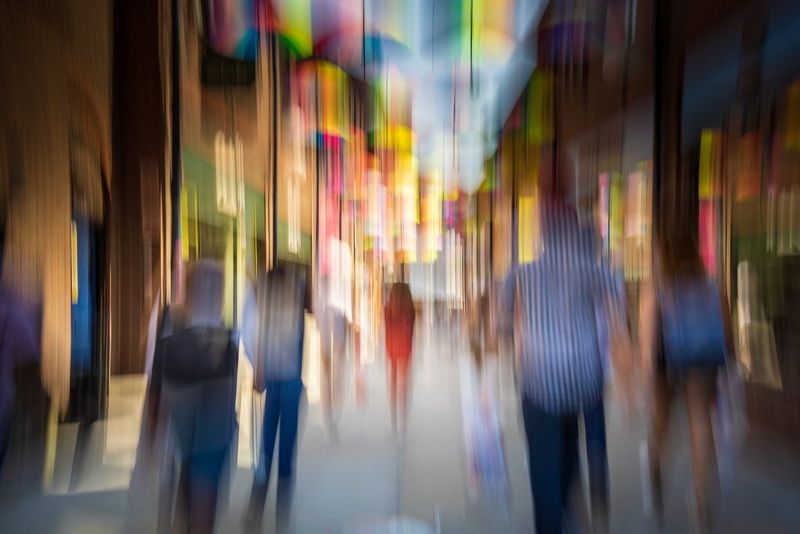 A street photography image with heavy vertical blur; pedestrians are visible amid streaks of pink, green, and yellow lights.