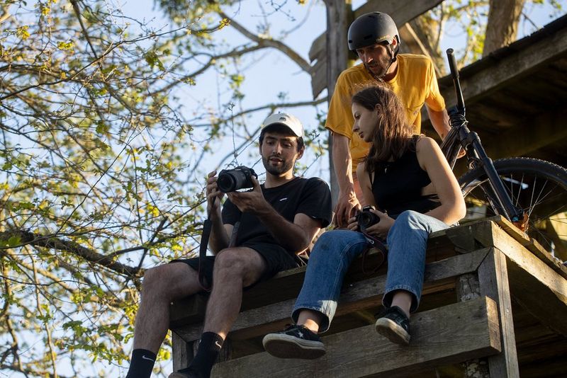 Trojica na leseni ploščadi, ki si ogleduje fotografije v fotoaparatu Canon EOS RP. Dva od trojice sedita, tretji s čelado pa stoji ob gorskem kolesu.