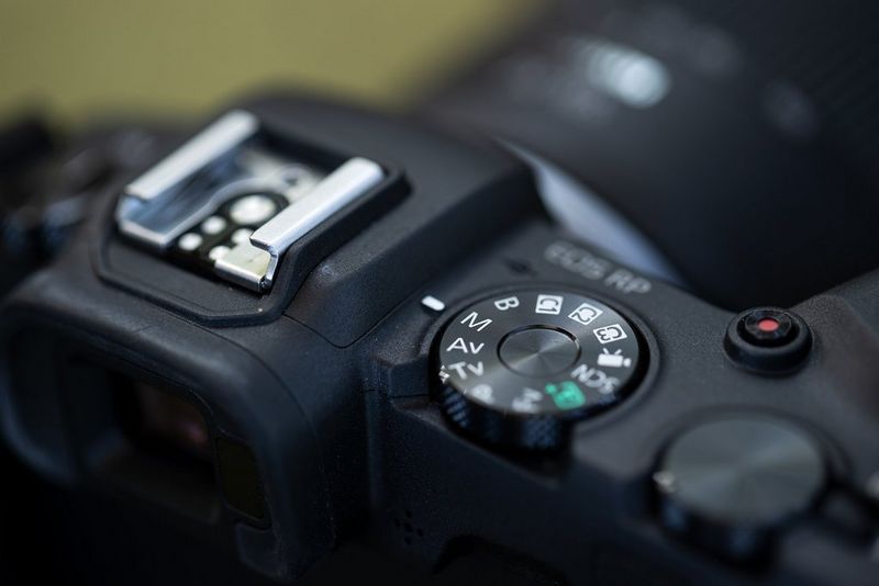 Slika zgornjega dela fotoaparata Canon EOS RP z izostrenim vrtljivim gumbom za izbiro načina fotografiranja.