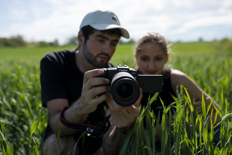 Canonov Ambasador Jean-Baptiste Liautard čepi na travi poleg Deborah in ji kaže vrtljivi zaslon na fotoaparatu Canon EOS RP.