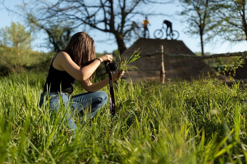 Mladenka v visoki travi, ki pred fotoaparatom drži šop trave, medtem ko sta v daljavi dva kolesarja na rampi.