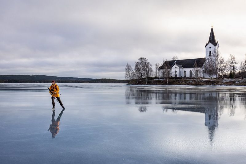 Uomo con giacca gialla che pattina su un lago ghiacciato. Nel ghiaccio, si vede il riflesso di una chiesa bianca.