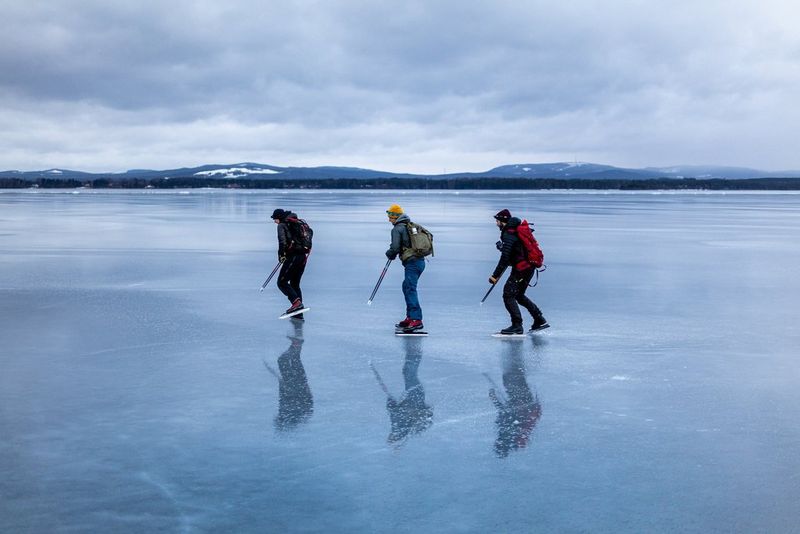 Tre pattinatori con i loro zaini in spalla attraversano un vasto lago ghiacciato. Cime innevate sono visibili in lontananza.