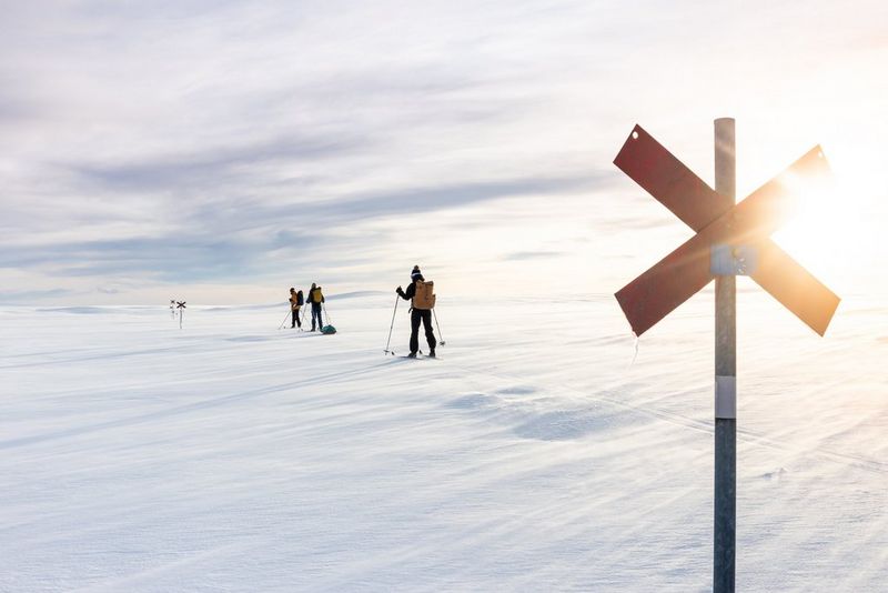 Tre fondisti attraversano un paesaggio innevato. Raggi di sole che illuminano un palo segnaletico dietro di loro.