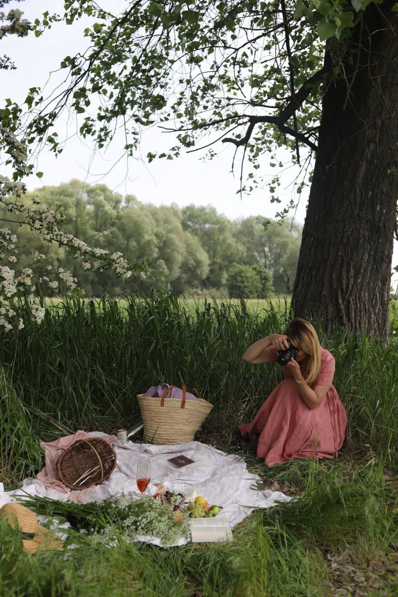 Žena u dugačkoj ružičastoj haljini sjedi sa strane bijele deke prekrivene cvijećem, hranom i pićem za piknik. Nalaze se na malom travnatom proplanku uz veliko stablo.