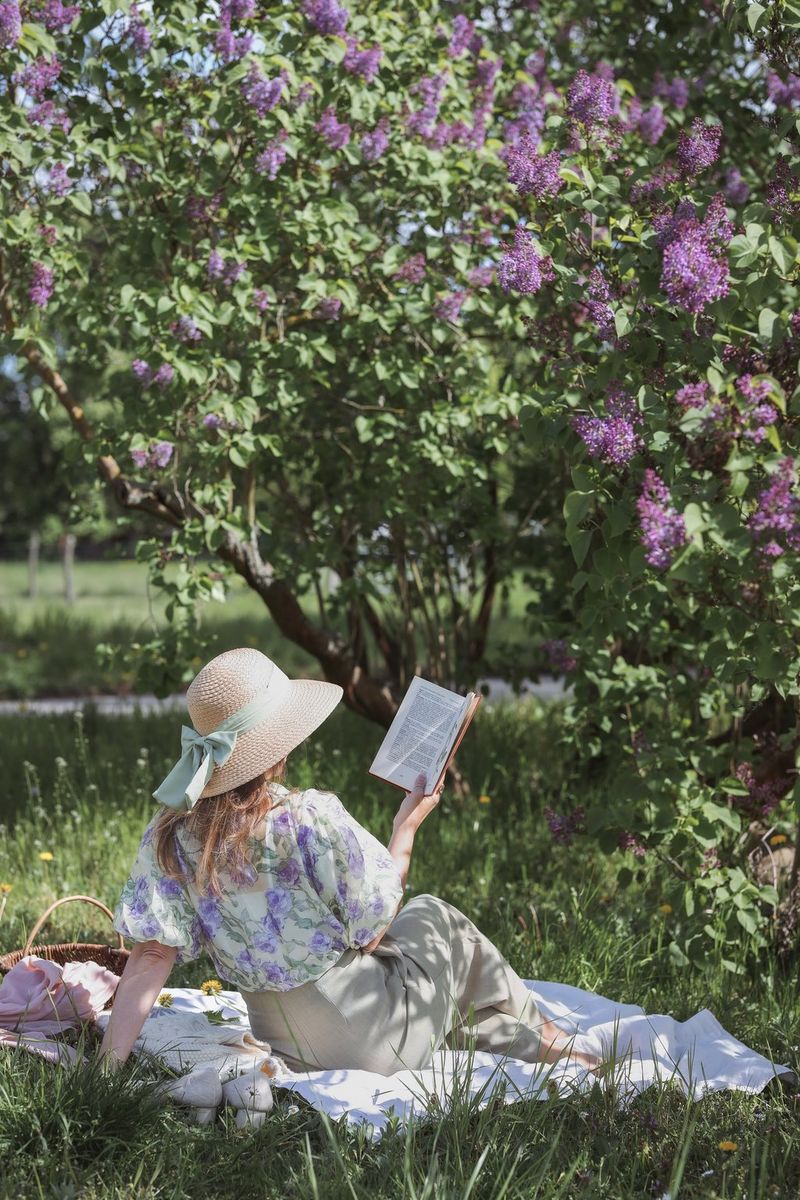 Žena sa slamnatim šeširom sjedi na deki za piknik i čita knjigu ispod stabla s ljubičastim cvjetovima.