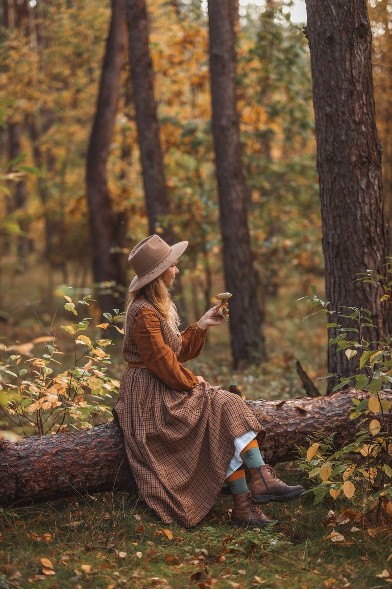 Žena s gljivom u ruci okrenuta udesno sjedi na deblu palog stabla u šumi.
