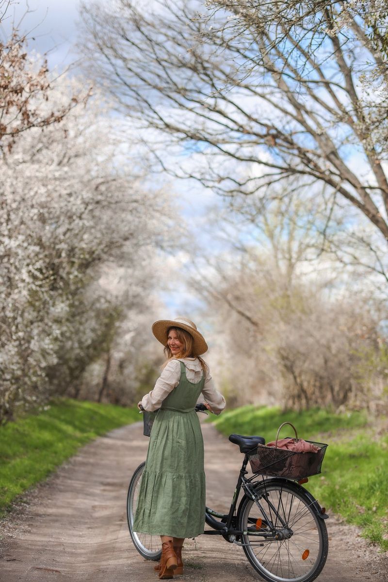 Na stazi sa stablima s obje strane žena drži upravljač bicikla te gleda unatrag i smiješi se fotoaparatu.