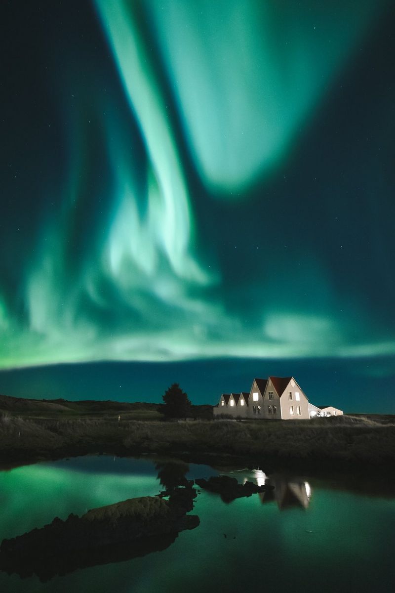 Dramatična zelena svjetla aurore borealis iznad kuće s osvijetljenim prozorima pokraj velike vodene površine.