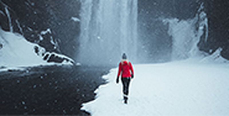 A woman dressed in a bright red winter jacket walks towards the Skógafoss waterfall in Iceland, as heavy snow falls around her.