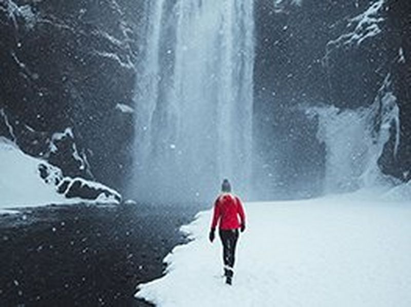 A woman dressed in a bright red winter jacket walks towards the Skógafoss waterfall in Iceland, as heavy snow falls around her.