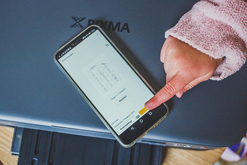 The hand of a small child touching a smartphone that is on top of a Canon PIXMA TS5340 printer.