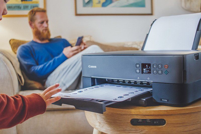 A child takes a printout from the paper tray of a Canon PIXMA TS5340 printer. A man sits on a sofa in the background looking at a smartphone.