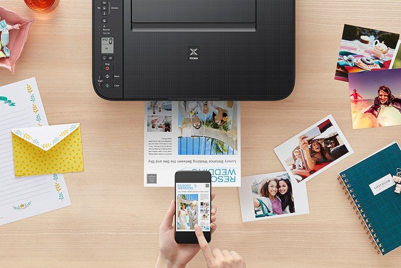 A Canon PIXMA TS3340 printer on a desk. Printouts, photos and a notebook are on the desk beside it.